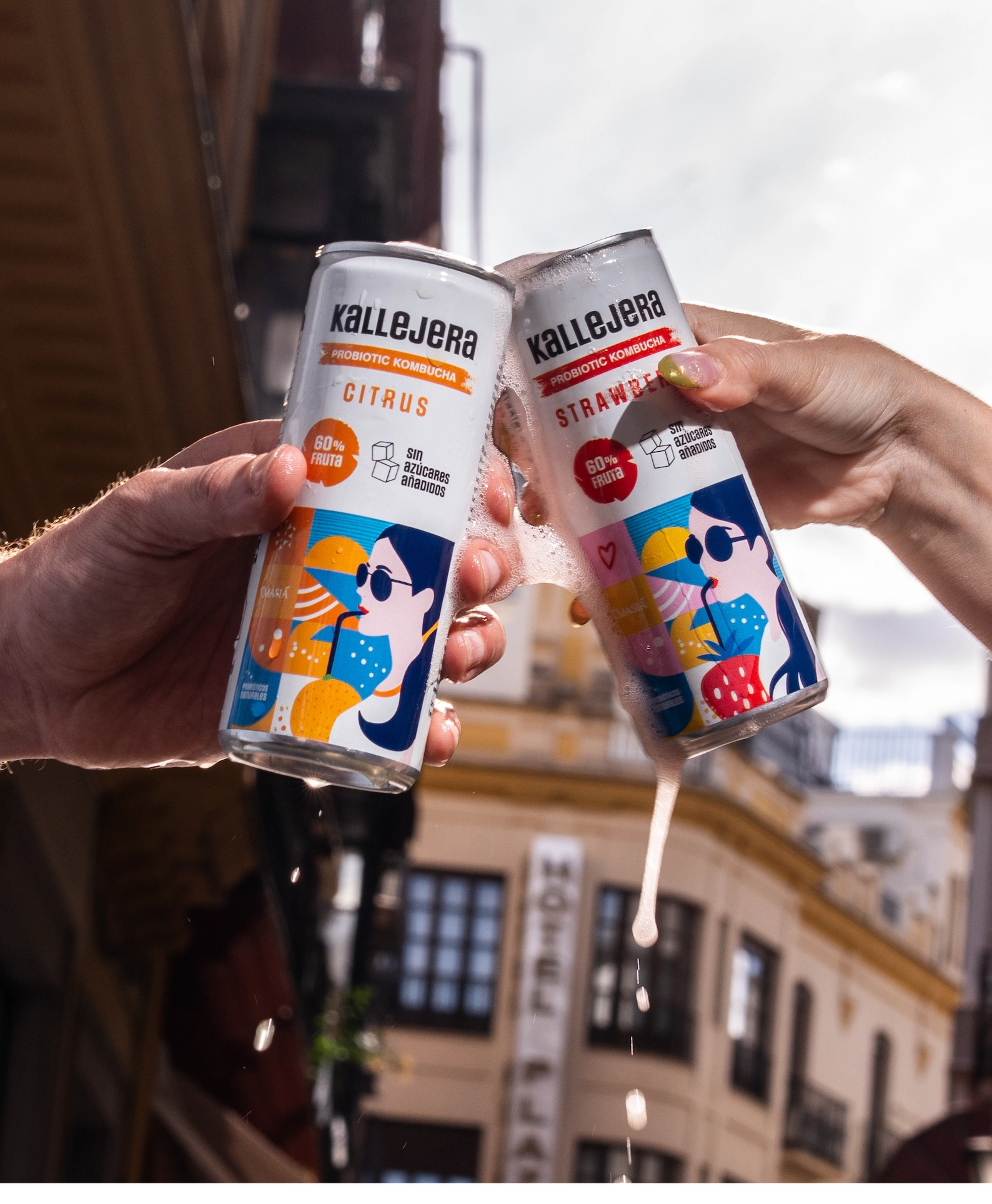 Dos manos brindando con latas de kombucha Kallejera salpicando.