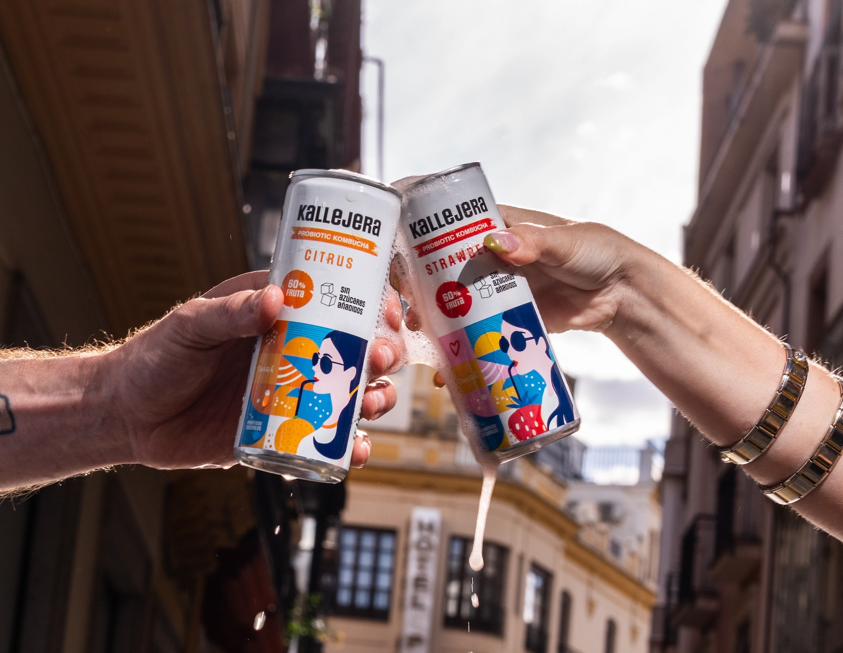 Dos manos brindando con latas de kombucha Kallejera en exteriores.